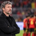 Een lid van de technische staf van België staat langs de zijlijn terwijl spelers op de achtergrond zichtbaar zijn.