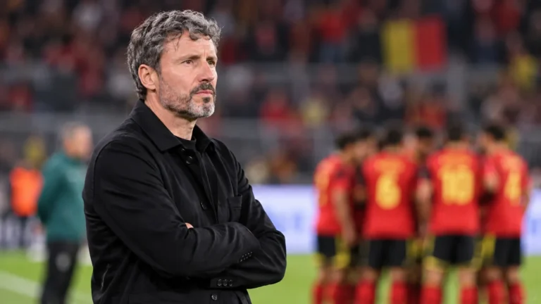 Een lid van de technische staf van België staat langs de zijlijn terwijl spelers op de achtergrond zichtbaar zijn.