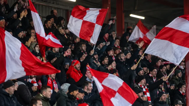 Een groep KV Kortrijk-supporters zwaait met rood-witte vlaggen op de tribune.