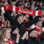 Een collectieve groep Arsenal-supporters viert opgelucht in de thuistribune met een rood-witte sjaal.
