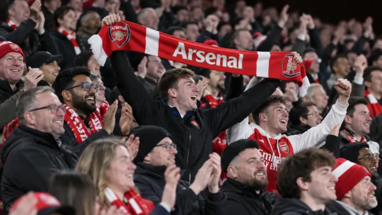 Een collectieve groep Arsenal-supporters viert opgelucht in de thuistribune met een rood-witte sjaal.