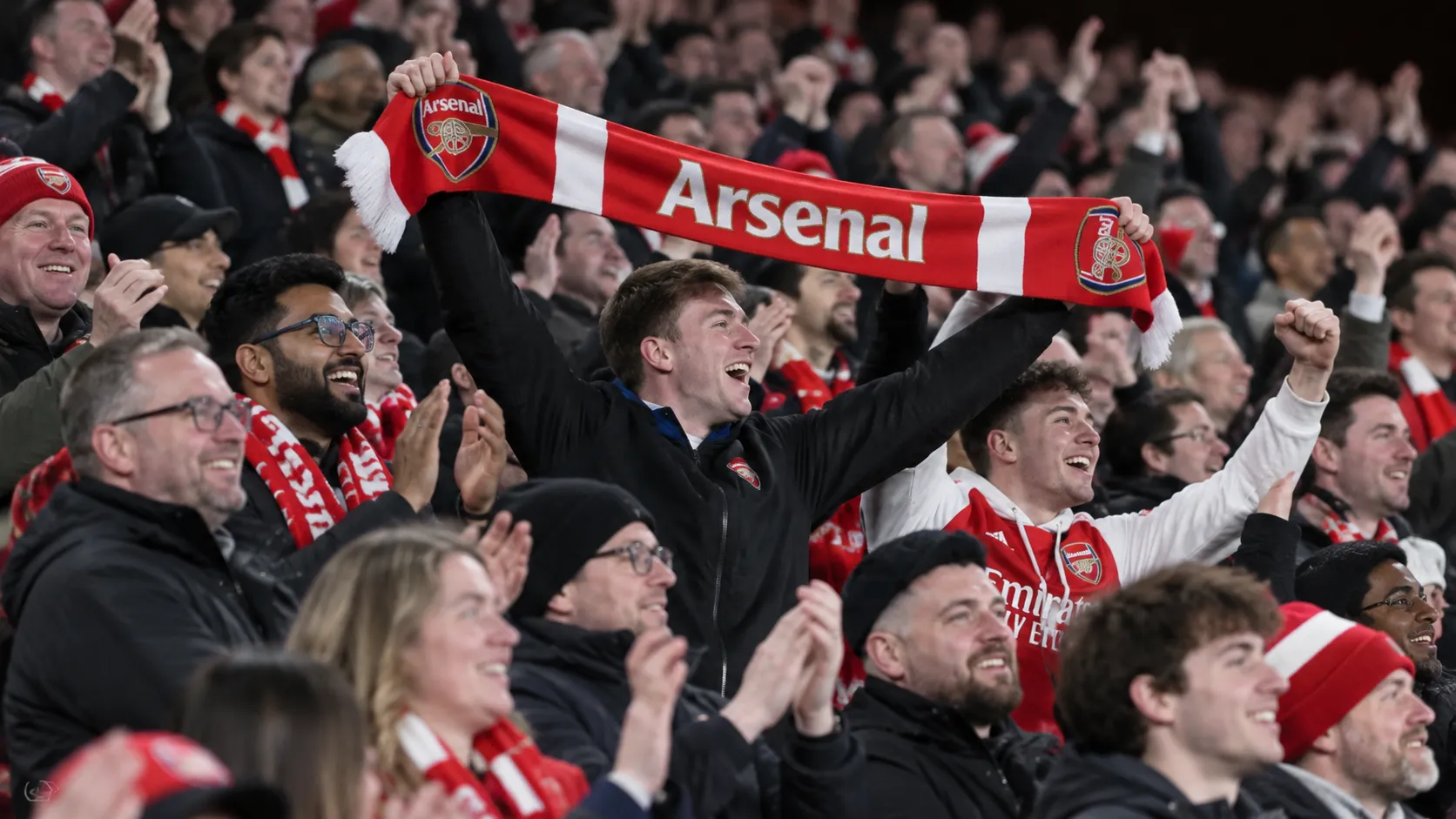 Een collectieve groep Arsenal-supporters viert opgelucht in de thuistribune met een rood-witte sjaal.