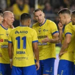 Een groep STVV-spelers in geel en blauw verzamelt zich op het veld tijdens een neutraal spelmoment.