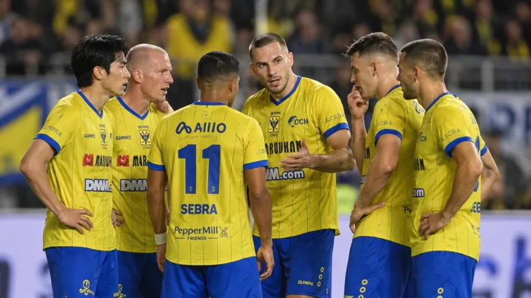 Een groep STVV-spelers in geel en blauw verzamelt zich op het veld tijdens een neutraal spelmoment.