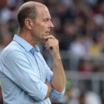 Een coach in een lichtblauw hemd kijkt geconcentreerd toe langs de zijlijn van een voetbalstadion.