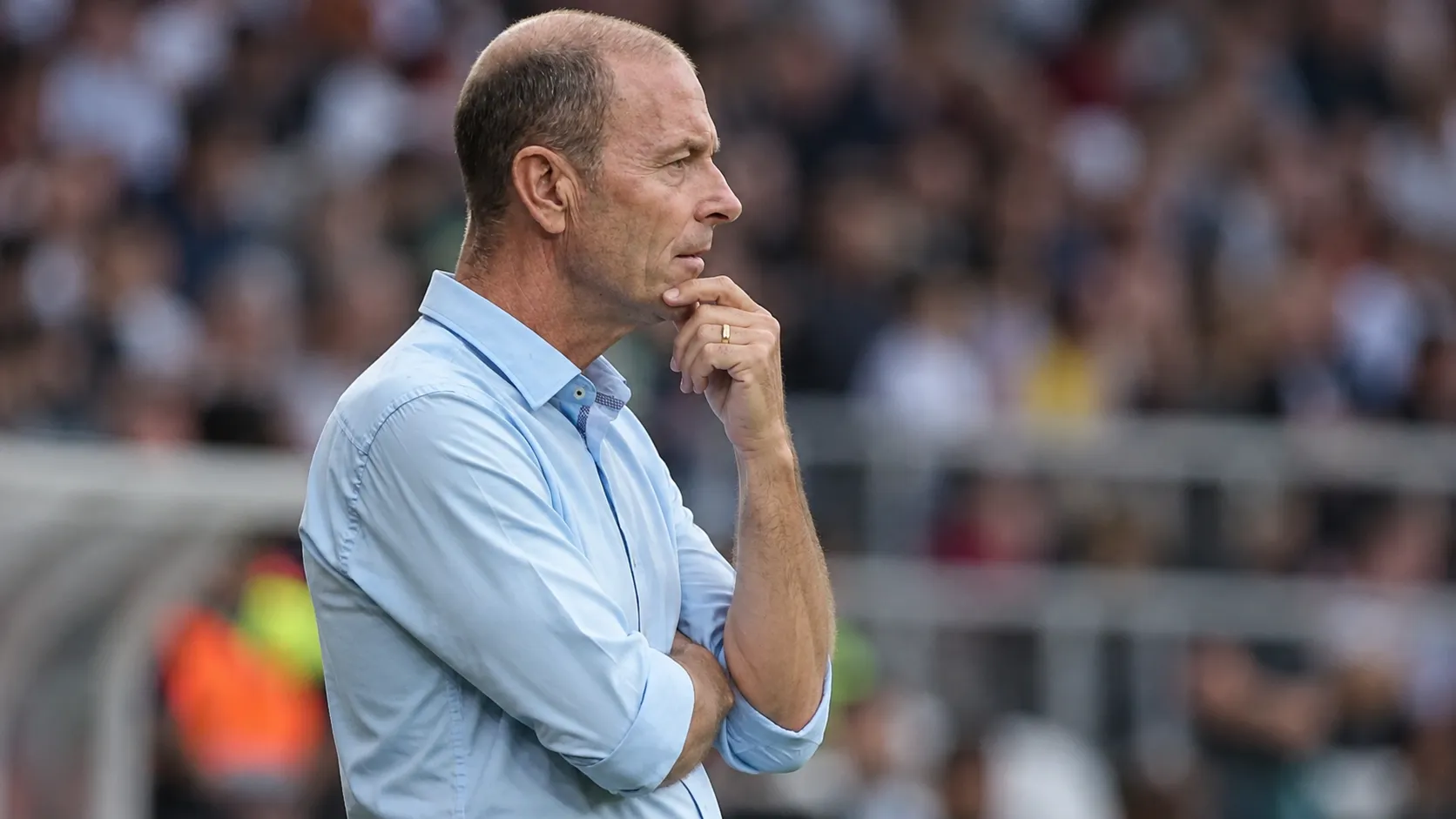 Een coach in een lichtblauw hemd kijkt geconcentreerd toe langs de zijlijn van een voetbalstadion.