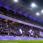 Paars-witte supporters met vlaggen in een verlichte stadiontribune van RSC Anderlecht.