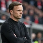 Een coach in een donkere trui langs de zijlijn van een stadion.