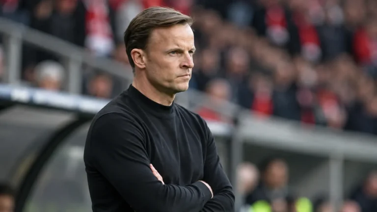 Een coach in een donkere trui langs de zijlijn van een stadion.