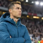 Cristiano Ronaldo in een blauwe gewatteerde jas naast de zijlijn van een voetbalstadion.