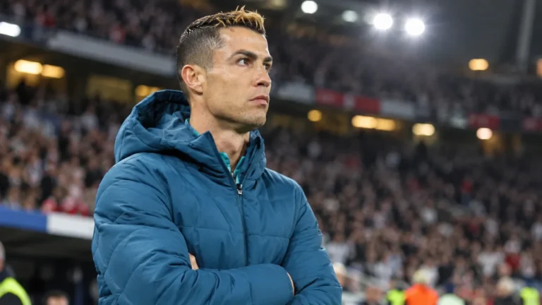 Cristiano Ronaldo in een blauwe gewatteerde jas naast de zijlijn van een voetbalstadion.