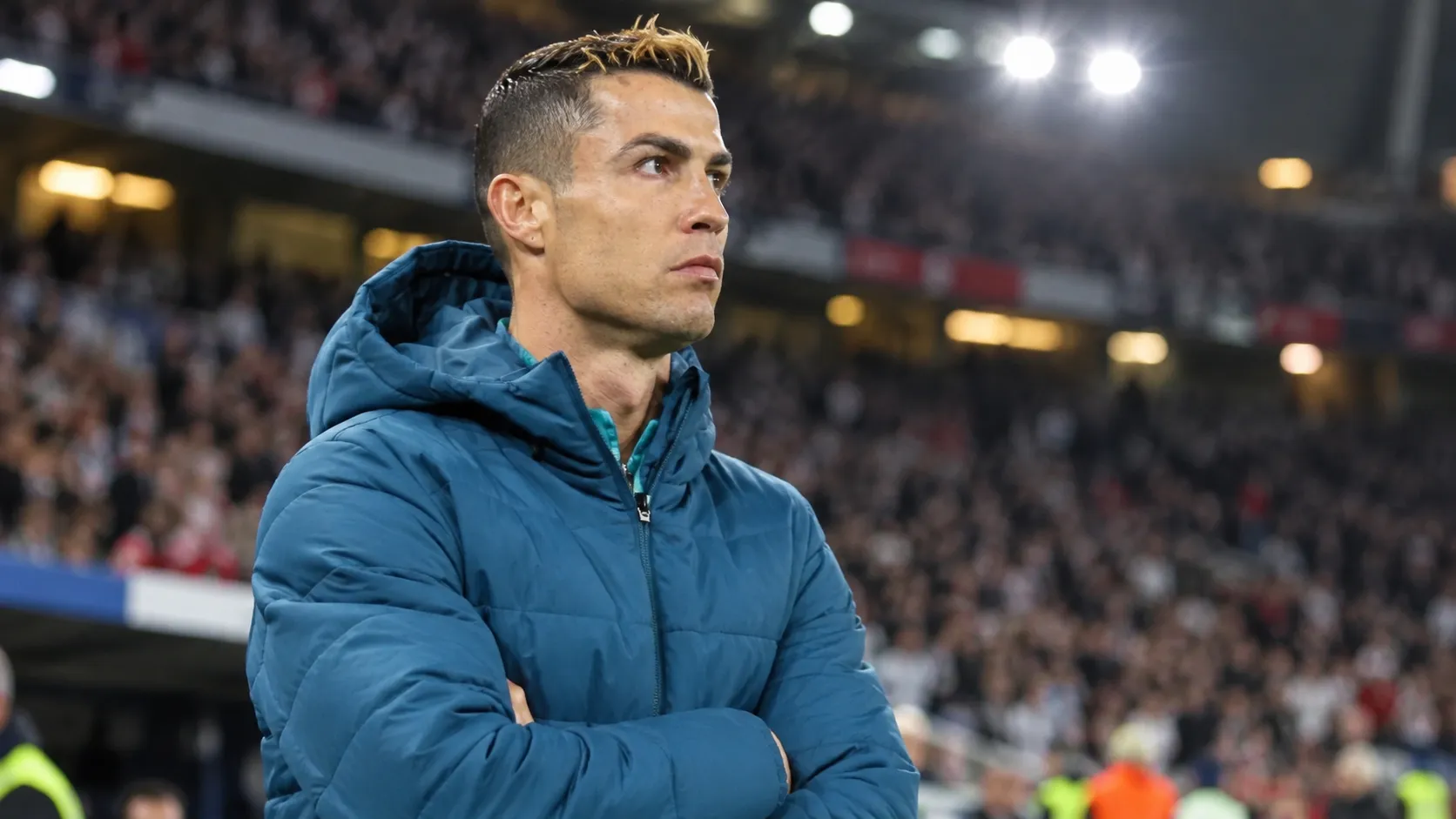 Cristiano Ronaldo in een blauwe gewatteerde jas naast de zijlijn van een voetbalstadion.