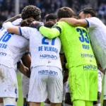 Een groep voetballers in witte shirts met blauwe accenten vormt na de wedstrijd een huddle.