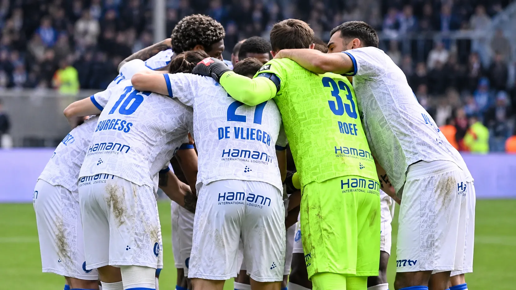 Een groep voetballers in witte shirts met blauwe accenten vormt na de wedstrijd een huddle.
