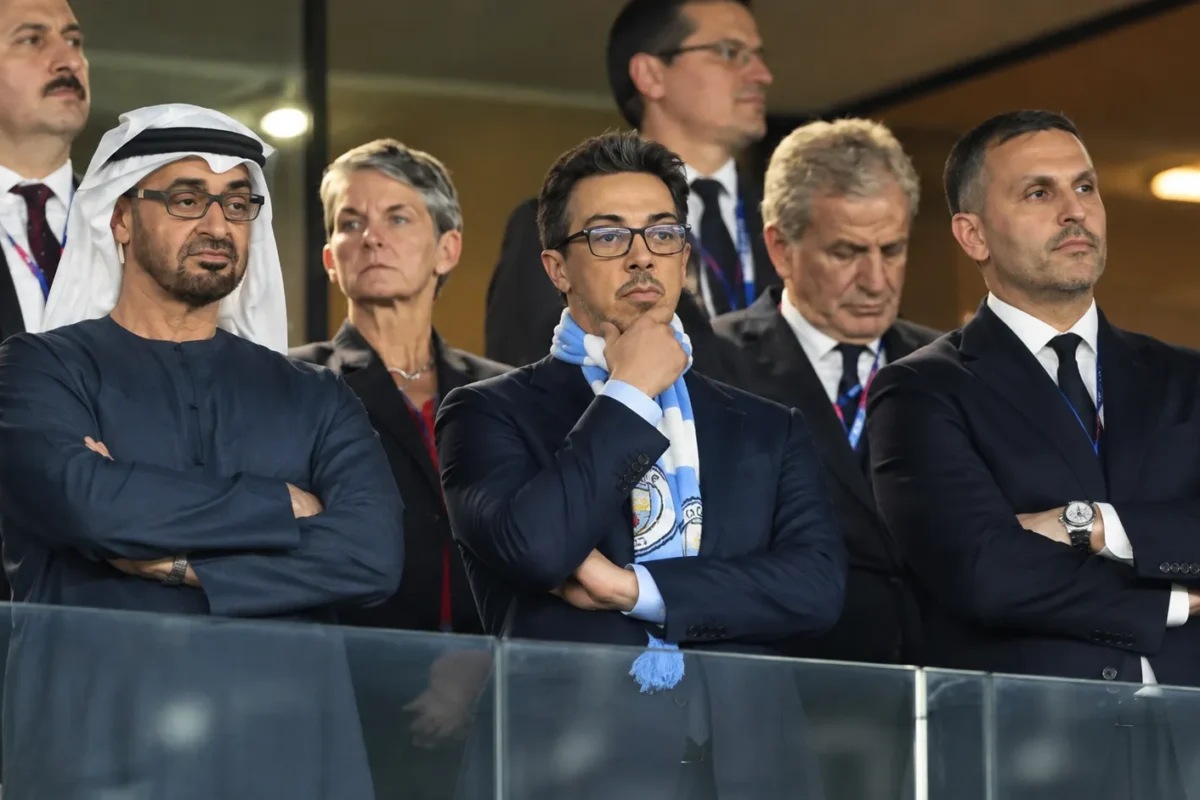 Een groep formeel geklede aanwezigen in een stadionloge, met een lichtblauwe supporterssjaal op de voorgrond.