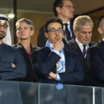 Een groep formeel geklede aanwezigen in een stadionloge, met een lichtblauwe supporterssjaal op de voorgrond.