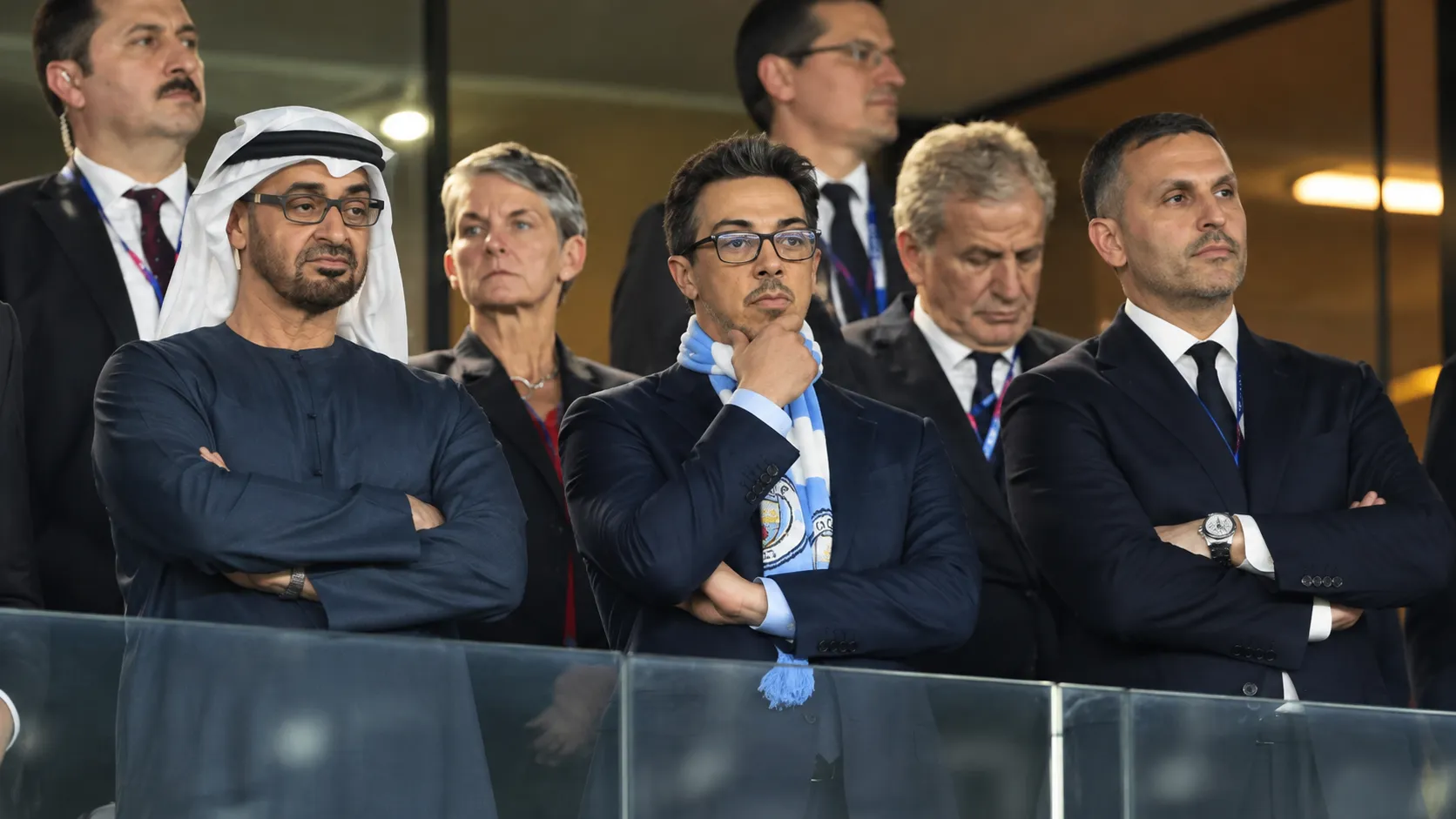 Een groep formeel geklede aanwezigen in een stadionloge, met een lichtblauwe supporterssjaal op de voorgrond.