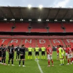 Spelers en scheidsrechters staan opgesteld voor lege rode tribunes in Sclessin.