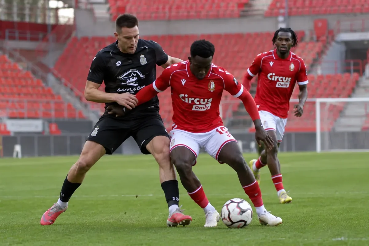 Voetballers in rode en zwarte wedstrijdtenues strijden om de bal in een leeg stadion.