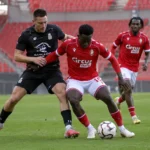 Voetballers in rode en zwarte wedstrijdtenues strijden om de bal in een leeg stadion.