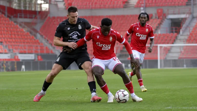 Voetballers in rode en zwarte wedstrijdtenues strijden om de bal in een leeg stadion.