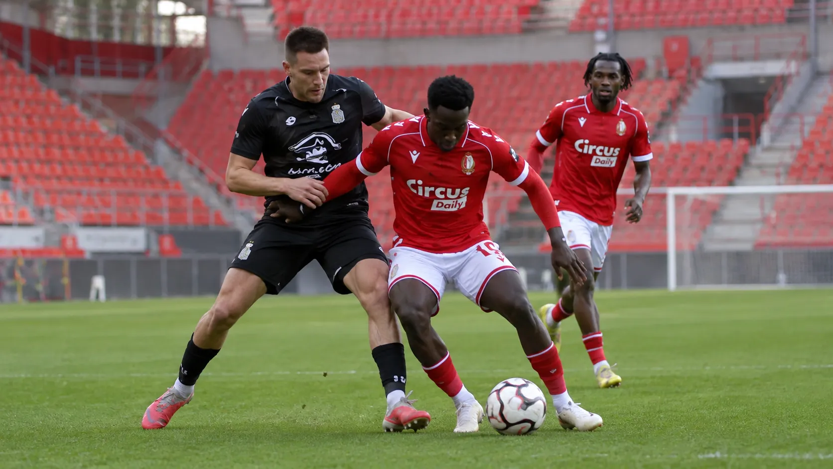 Voetballers in rode en zwarte wedstrijdtenues strijden om de bal in een leeg stadion.