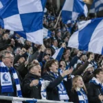 Een volle tribune met KAA Gent-supporters die blauw-witte vlaggen zwaaien.