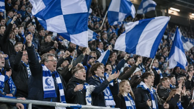 Een volle tribune met KAA Gent-supporters die blauw-witte vlaggen zwaaien.