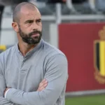 Steven Defour in een grijze trui naast het veld met een subtiel Belgisch nationaal teamembleem op de achtergrond.