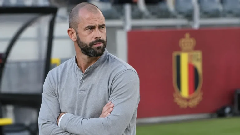 Steven Defour in een grijze trui naast het veld met een subtiel Belgisch nationaal teamembleem op de achtergrond.
