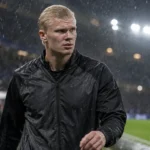Erling Haaland in een donkere jas aan de rand van een regenachtig stadionveld.