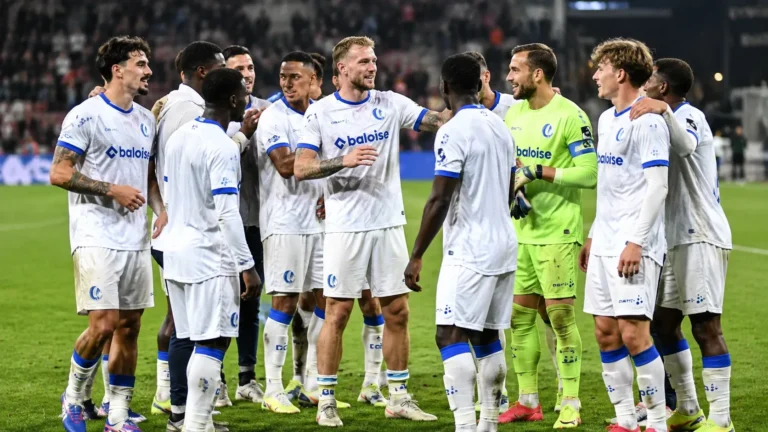 Een groep voetballers in witte tenues met blauwe accenten verzamelt op het gras na de wedstrijd.