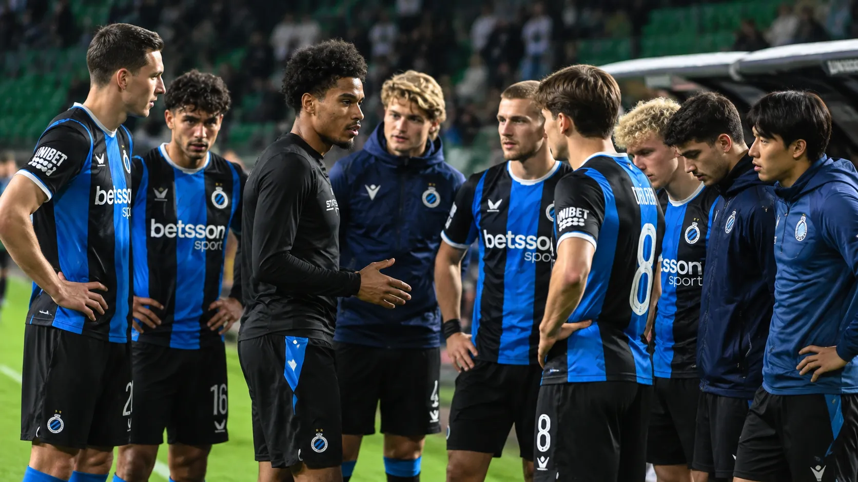 Een groep spelers in zwart-blauwe clubtenues verzamelt zich langs het veld voor een Europese avondwedstrijd.