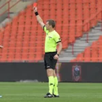 Scheidsrechter toont een rode kaart op het veld, met spelers in rood en zwart bij lege tribunes.