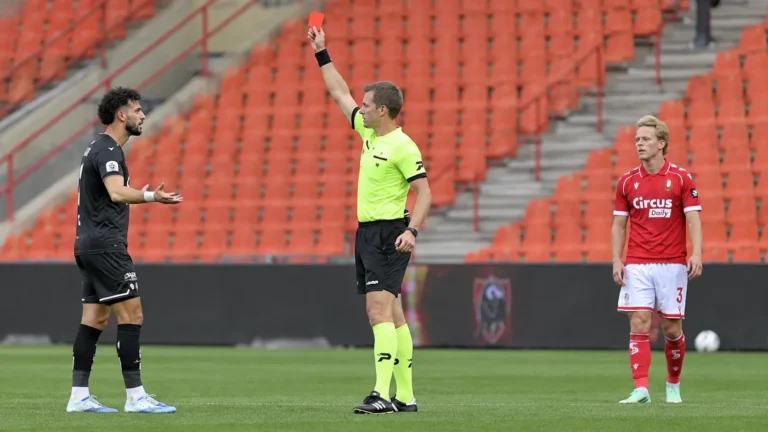 Scheidsrechter toont een rode kaart op het veld, met spelers in rood en zwart bij lege tribunes.