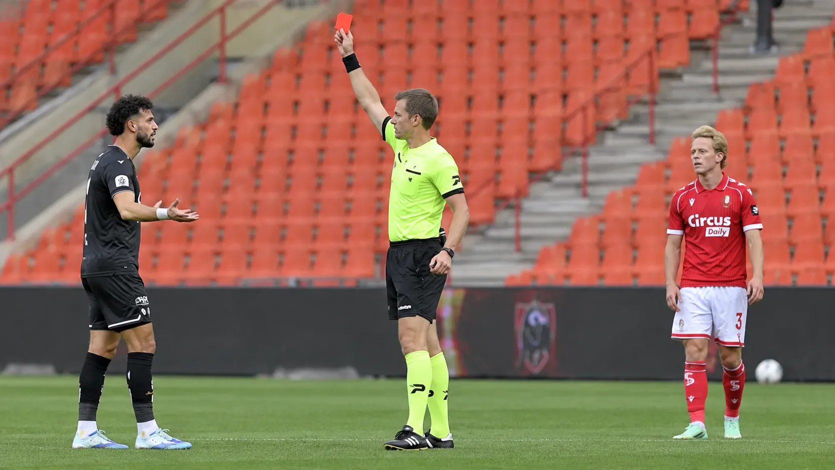 Scheidsrechter toont een rode kaart op het veld, met spelers in rood en zwart bij lege tribunes.