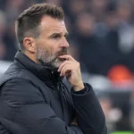 Een coach in donkere jas kijkt aandachtig vanaf de zijlijn van een stadion.