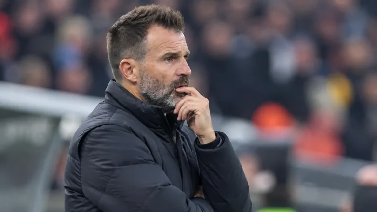Een coach in donkere jas kijkt aandachtig vanaf de zijlijn van een stadion.