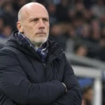 Een trainer in een donkere jas observeert het veld vanuit de technische zone van een stadion.