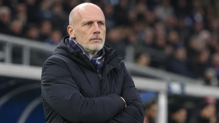 Een trainer in een donkere jas observeert het veld vanuit de technische zone van een stadion.