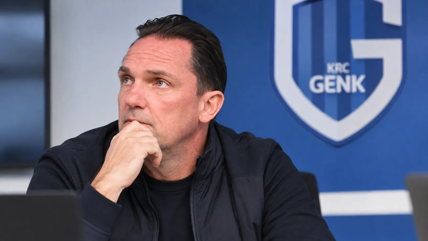 Een KRC Genk-staflid in donkere clubkleding tijdens een intern overleg in een vergaderruimte.