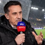 Gary Neville spreekt met een microfoon aan een overdekte commentaarpositie bij een regenachtig stadion.