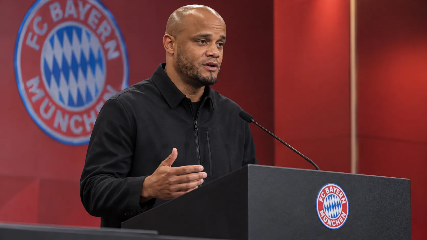 Vincent Kompany spreekt op een persmoment van Bayern München achter een spreekgestoelte.