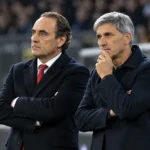 Twee coaches in donkere jassen naast het veld in een Belgisch voetbalstadion.