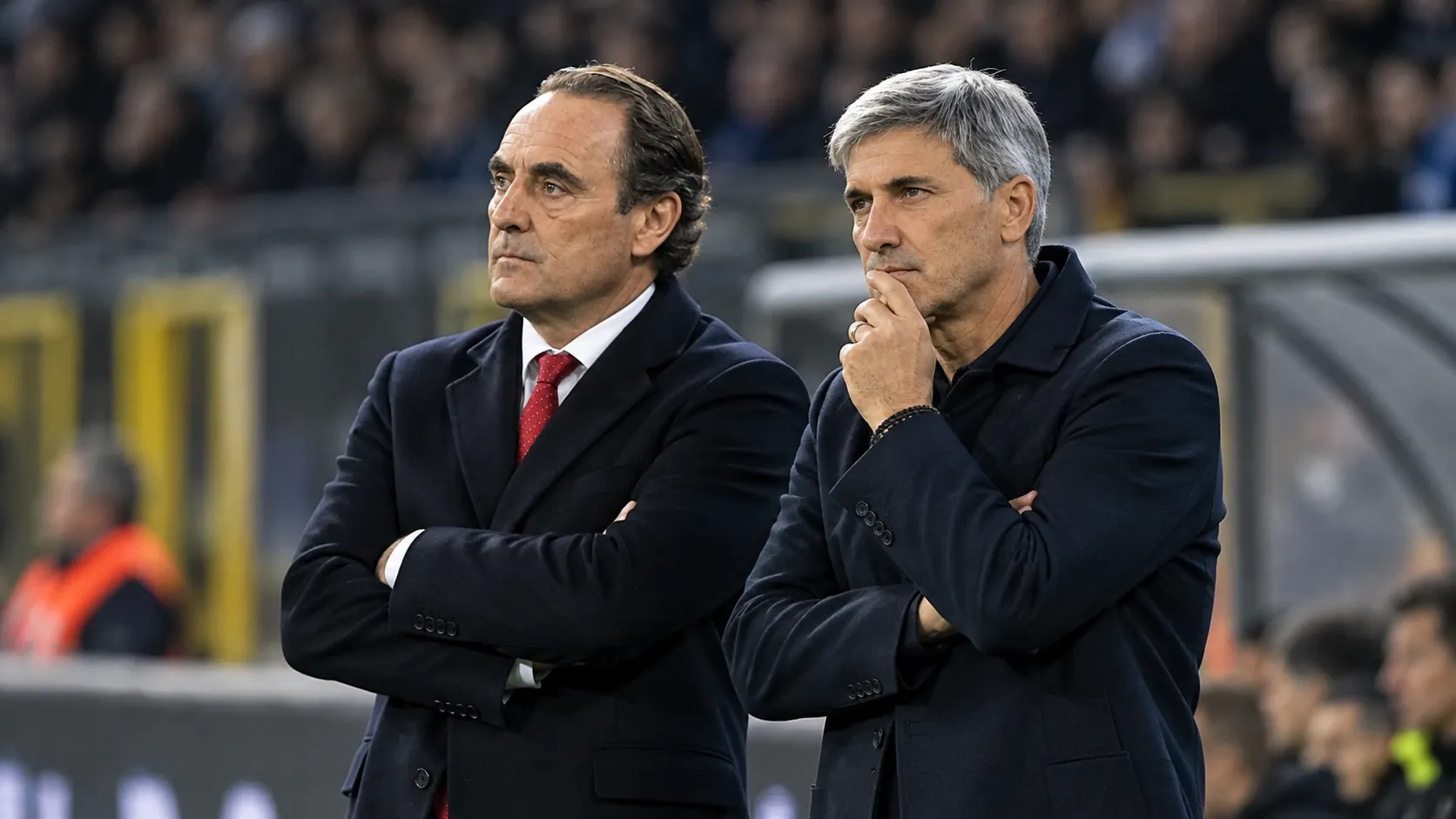 Twee coaches in donkere jassen naast het veld in een Belgisch voetbalstadion.