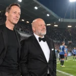 Twee niet-spelende clubvertegenwoordigers staan langs het veld in een verlicht stadion.