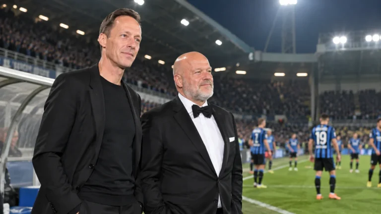 Twee niet-spelende clubvertegenwoordigers staan langs het veld in een verlicht stadion.