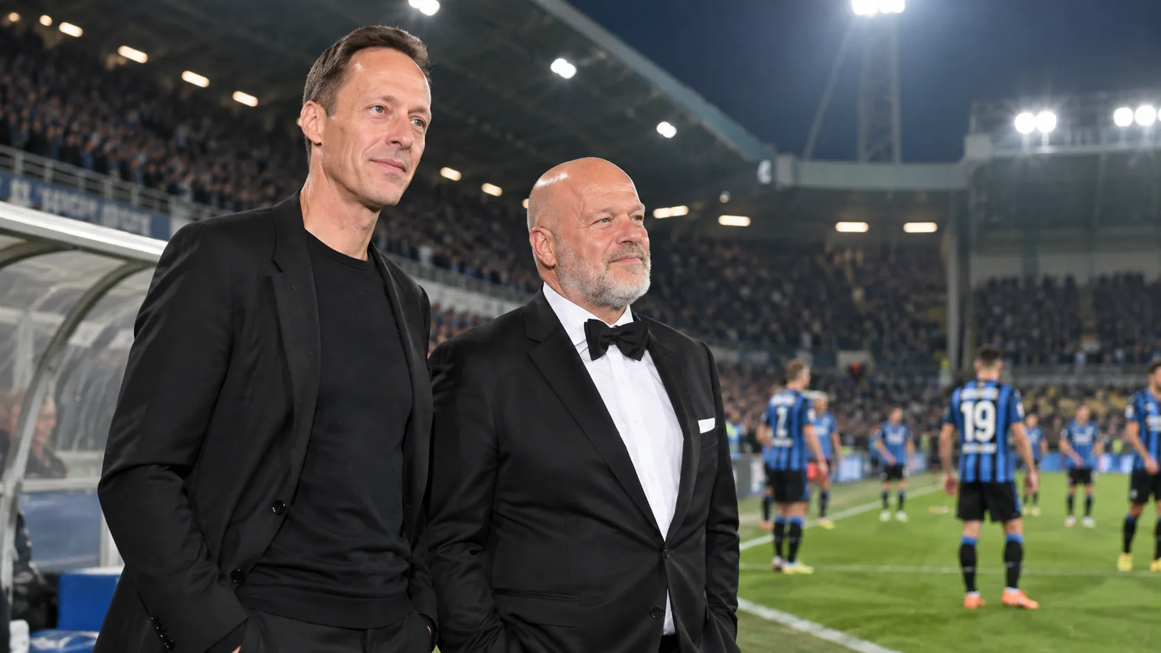 Twee niet-spelende clubvertegenwoordigers staan langs het veld in een verlicht stadion.