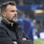 Een coach in een zwarte jas kijkt geconcentreerd vanaf de zijlijn.