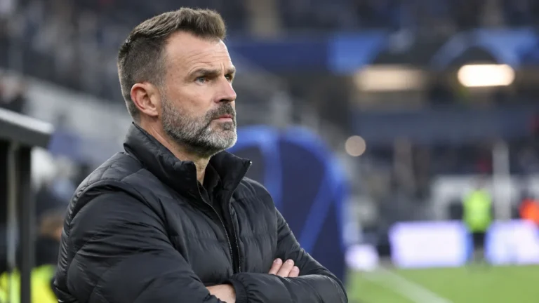 Een coach in een zwarte jas kijkt geconcentreerd vanaf de zijlijn.